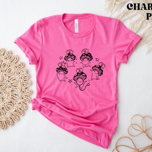 Op de afbeelding: Roze T-shirt met een zwarte afbeelding van cartoon-paddenstoelen die verpleegstersmutsen dragen. De tekst "Shroom Queens" is op het shirt gedrukt.