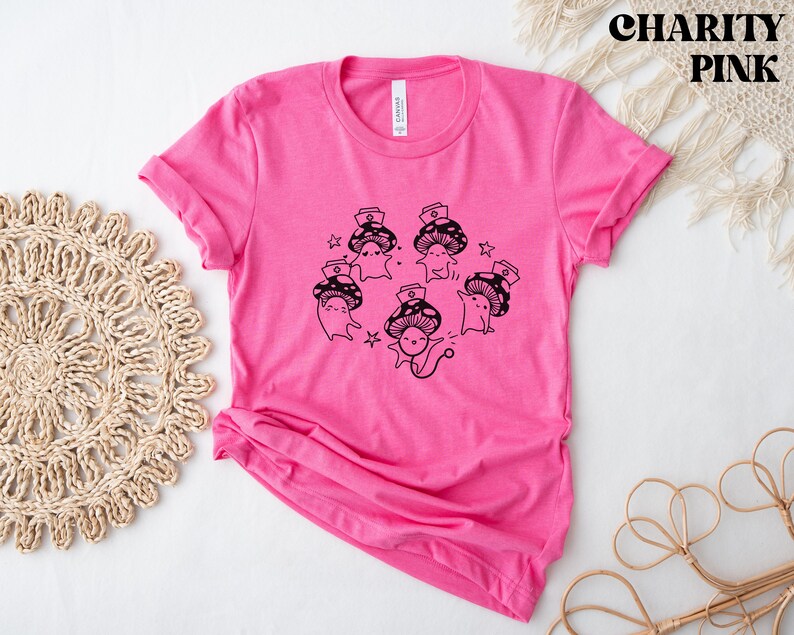 Op de afbeelding: Roze T-shirt met een zwarte afbeelding van cartoon-paddenstoelen die verpleegstersmutsen dragen. De tekst "Shroom Queens" is op het shirt gedrukt.