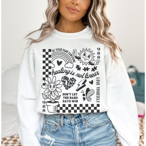 May include: White cropped sweatshirt with black graphic print featuring a flower, rainbow, sun, peace sign, and text that reads "Be your own sunshine", "healing is not linear", "Find joy in the journey", "Don't let the hard days win", "Love yourself", and "988".