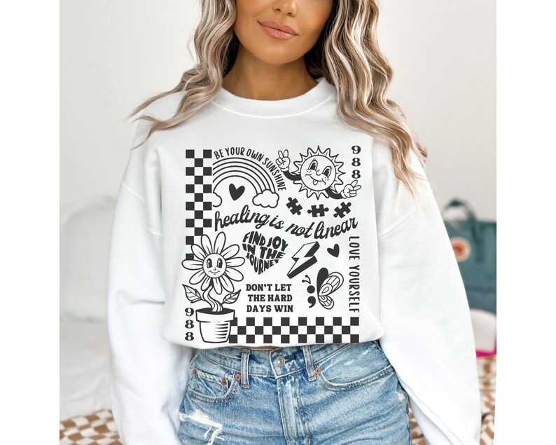 May include: White cropped sweatshirt with black graphic print featuring a flower, rainbow, sun, peace sign, and text that reads "Be your own sunshine", "healing is not linear", "Find joy in the journey", "Don't let the hard days win", "Love yourself", and "988".