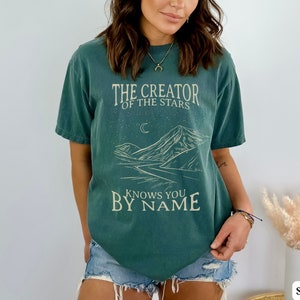 May include: A teal green t-shirt with a mountain landscape graphic and the text "The Creator of the Stars Knows You By Name".