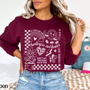 May include: A maroon cropped sweatshirt with a white graphic print featuring a sun, a flower, a rainbow, and the text "Be your own sunshine", "Healing is not linear", "Find joy in the journey", "Don't let the hard days win", "Love yourself", and "988".