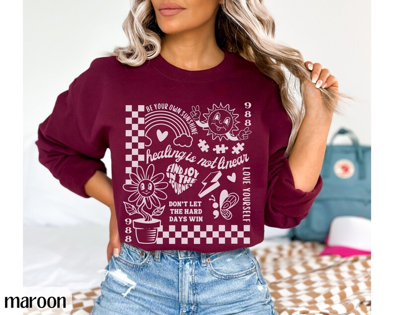 May include: A maroon cropped sweatshirt with a white graphic print featuring a sun, a flower, a rainbow, and the text "Be your own sunshine", "Healing is not linear", "Find joy in the journey", "Don't let the hard days win", "Love yourself", and "988".