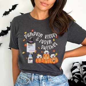 May include: A dark gray t-shirt with the text "Pumpkin Kisses & Zosyn Wishes" and a graphic of three pumpkins wearing nurse hats. One pumpkin has a witch's hat on top of the nurse hat. A medical IV bag and a bottle of medication are in the background.