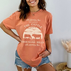 May include: A coral colored t-shirt with a white graphic design that says "He owns the cattle on a thousand hills, Psalm 50". The graphic also includes a silhouette of cattle.