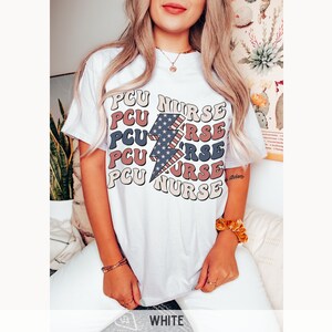 K&ouml;nnte beinhalten: Wei&szlig;es T-Shirt mit einem Grafikdruck eines Blitzes mit einem amerikanischen Flaggenmuster und dem Text "PCU NURSE", der in einem gestapelten Muster wiederholt wird.