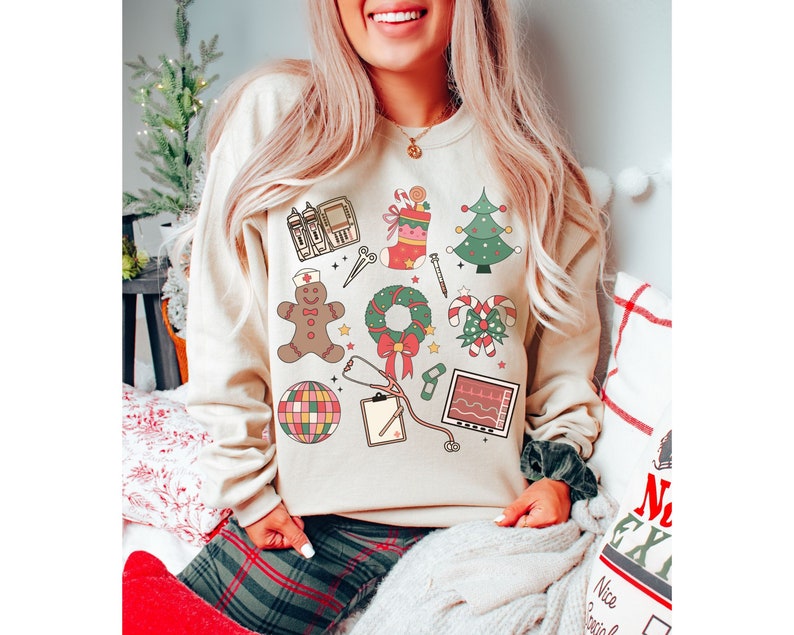 May include: A cream-colored sweatshirt with a festive Christmas design featuring a gingerbread person, a candy cane, a wreath, a stocking, a stethoscope, a heart monitor, a bandage, a Christmas tree, and a disco ball.