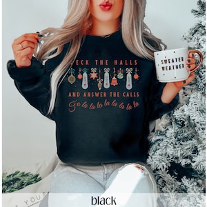 Może przedstawiać: Czarny świąteczny sweter z napisem "Deck the halls and answer the calls" i rzędem świątecznych ozd&oacute;b wiszących z napisu. Napis "fa la la la la la la la la" znajduje się pod ozdobami.