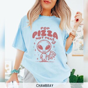 May include: A light blue t-shirt with a graphic of an alien holding a slice of pizza. The text on the shirt reads "Pop Pizza Not Pills".