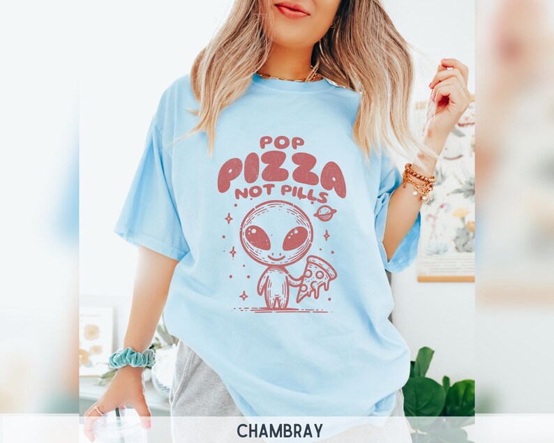 May include: A light blue t-shirt with a graphic of an alien holding a slice of pizza. The text on the shirt reads "Pop Pizza Not Pills".
