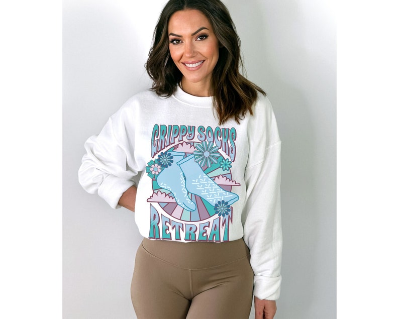 May include: White sweatshirt with a graphic of blue socks with a white snowflake pattern, surrounded by flowers and a rainbow. The text "Grippy Socks Retreat" is printed on the sweatshirt.