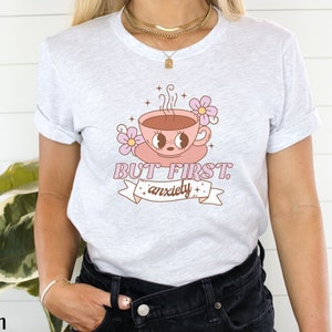 May include: A white t-shirt with a pink coffee cup with eyes and a pink flower. The text on the shirt reads "BUT FIRST, anxiety".