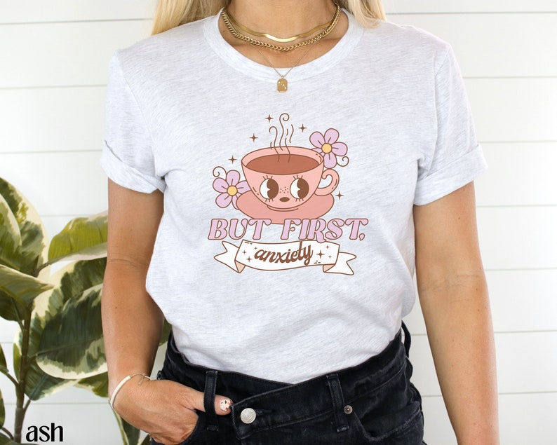 May include: A white t-shirt with a pink coffee cup with eyes and a pink flower. The text on the shirt reads "BUT FIRST, anxiety".