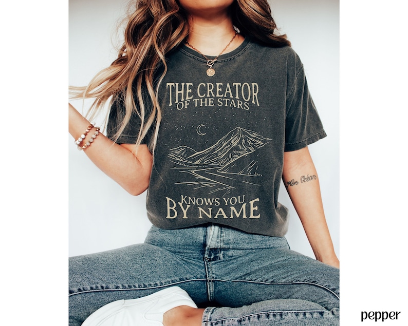May include: A dark grey t-shirt with a graphic design of a mountain range and the text "The Creator of the Stars Knows You By Name".