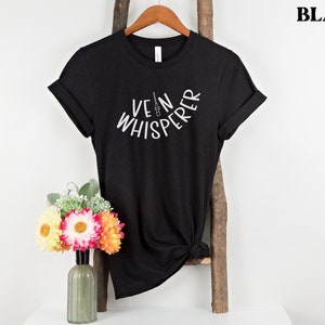 以下が含まれることがあります： 白い文字で「Vein Whisperer」とスタイリッシュな針の画像が書かれた黒いTシャツ