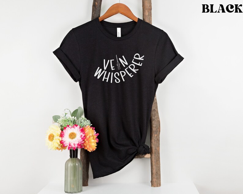 以下が含まれることがあります： 白い文字で「Vein Whisperer」とスタイリッシュな針の画像が書かれた黒いTシャツ