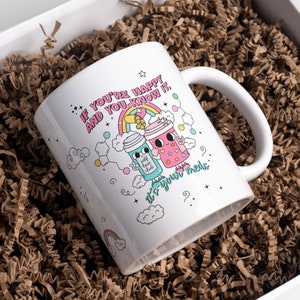 May include: White ceramic mug with a colourful illustration of two cartoon characters holding cups. The text on the mug reads "If you're happy and you know it, it's your meds."