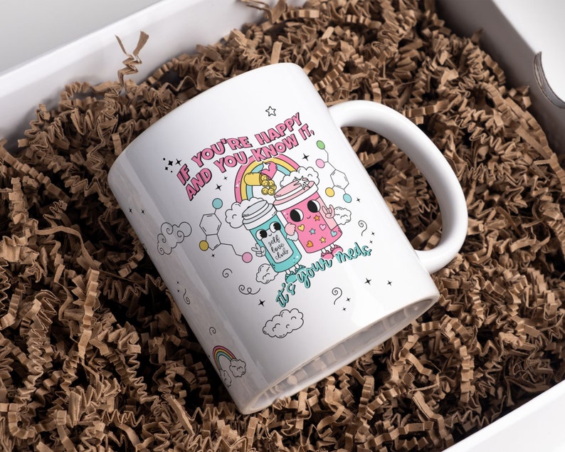 May include: White ceramic mug with a colourful illustration of two cartoon characters holding cups. The text on the mug reads "If you're happy and you know it, it's your meds."