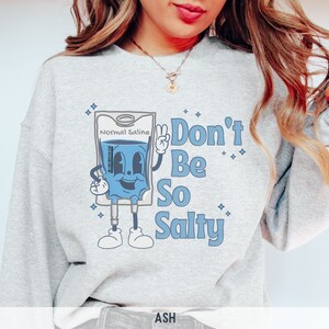 Op de afbeelding: Een grijs sweatshirt met een cartoonillustratie van een infuuszak met de tekst "Normal Saline" en de zin "Don't Be So Salty".
