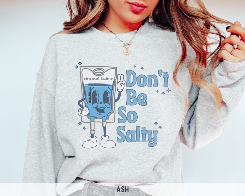 Op de afbeelding: Een grijs sweatshirt met een cartoonillustratie van een infuuszak met de tekst "Normal Saline" en de zin "Don't Be So Salty".