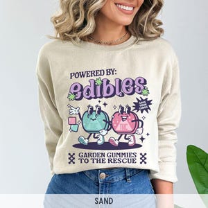 May include: A beige sweatshirt with a graphic print of two cartoon gummies. The gummies are green and pink and have faces. The text on the sweatshirt says "Powered by: edibles" and "Garden gummies to the rescue".