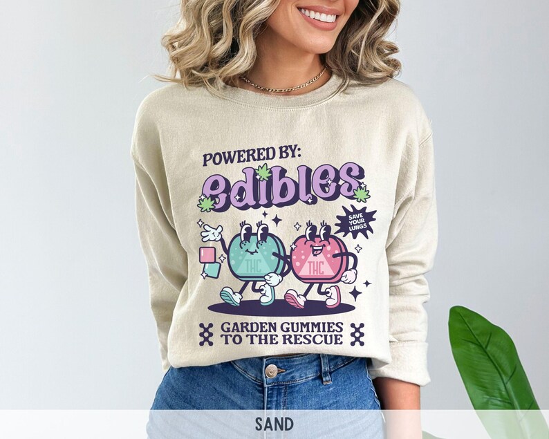 May include: A beige sweatshirt with a graphic print of two cartoon gummies. The gummies are green and pink and have faces. The text on the sweatshirt says "Powered by: edibles" and "Garden gummies to the rescue".