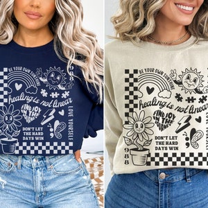 May include: Two navy blue and beige sweatshirts with a graphic print of a black and white checkered background with a rainbow, a sun, a flower, a heart, a lightning bolt, a butterfly, and the text "BE YOUR OWN SUNSHINE", "healing is not linear", "FIND JOY IN THE JOURNEY", "DON'T LET THE HARD DAYS WIN", and "LOVE YOURSELF".