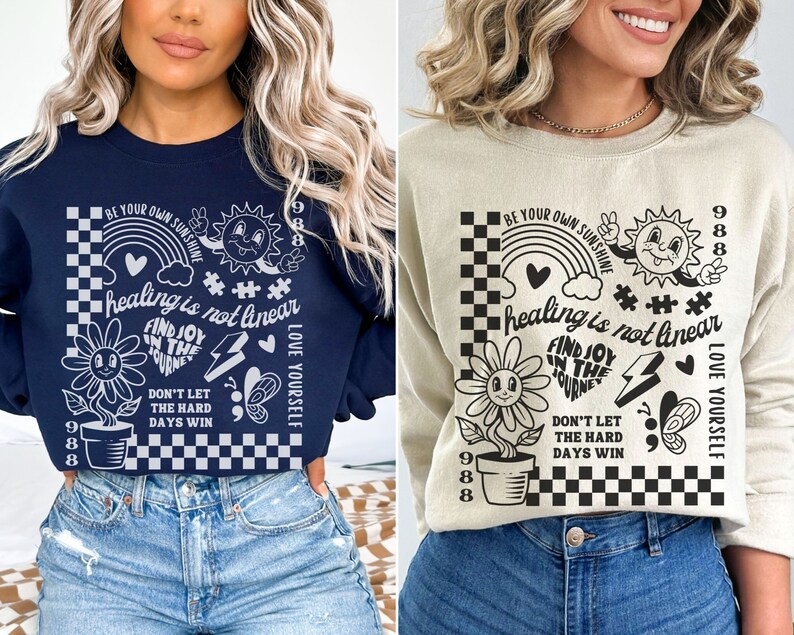May include: Two navy blue and beige sweatshirts with a graphic print of a black and white checkered background with a rainbow, a sun, a flower, a heart, a lightning bolt, a butterfly, and the text "BE YOUR OWN SUNSHINE", "healing is not linear", "FIND JOY IN THE JOURNEY", "DON'T LET THE HARD DAYS WIN", and "LOVE YOURSELF".
