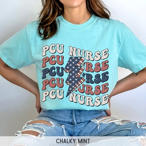 K&ouml;nnte beinhalten: Ein hellblaues T-Shirt mit einem Grafikdruck eines Blitzes mit rot-wei&szlig;-blauen Streifen. Der Text "PCU NURSE" wird in einer Retro-Schriftart um den Blitz herum wiederholt.
