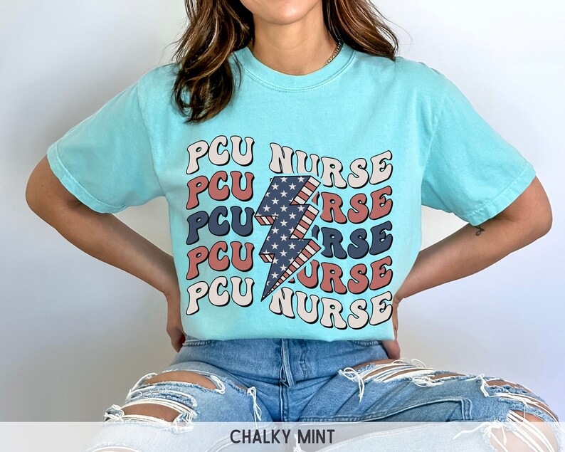 K&ouml;nnte beinhalten: Ein hellblaues T-Shirt mit einem Grafikdruck eines Blitzes mit rot-wei&szlig;-blauen Streifen. Der Text "PCU NURSE" wird in einer Retro-Schriftart um den Blitz herum wiederholt.