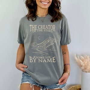 May include: A grey t-shirt with a graphic design of a mountain range, a crescent moon, and stars. The text "The Creator of the Stars Knows You By Name" is printed on the shirt.