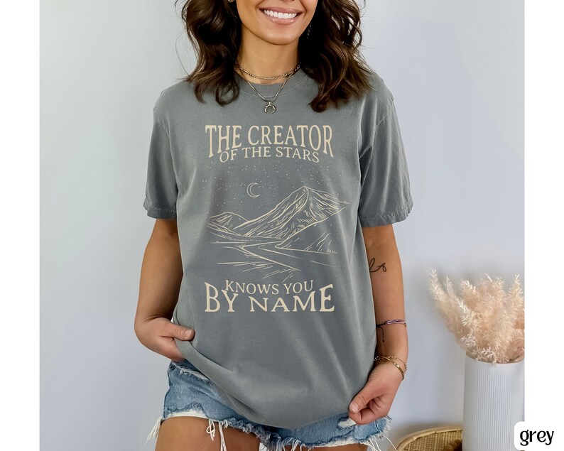 May include: A grey t-shirt with a graphic design of a mountain range, a crescent moon, and stars. The text "The Creator of the Stars Knows You By Name" is printed on the shirt.