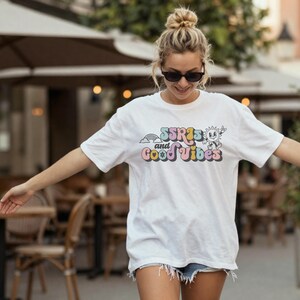 May include: White t-shirt with the text "SSRIs and Good Vibes" in colorful, retro-style lettering. The design includes a rainbow and a smiling sun character. The shirt is short-sleeved and has a relaxed fit.