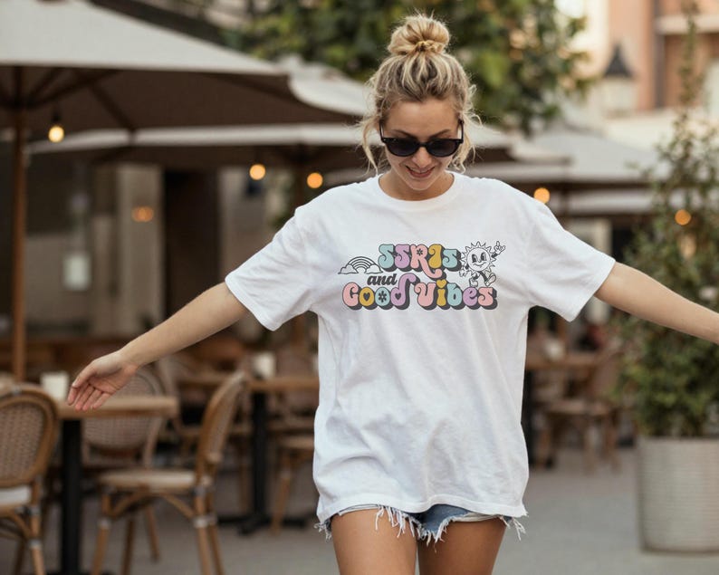 May include: White t-shirt with the text "SSRIs and Good Vibes" in colorful, retro-style lettering. The design includes a rainbow and a smiling sun character. The shirt is short-sleeved and has a relaxed fit.