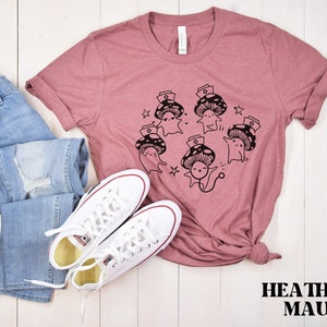 Op de afbeelding: Een heide-paarse t-shirt met een afbeelding van schattige cartoon-paddenstoelen die verpleegstersmutsen dragen. De paddenstoelen zijn in een cirkel gerangschikt met sterren eromheen. De tekst "HEATHER MAUVE" staat onderaan de afbeelding.