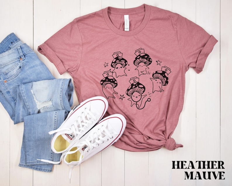 Op de afbeelding: Een heide-paarse t-shirt met een afbeelding van schattige cartoon-paddenstoelen die verpleegstersmutsen dragen. De paddenstoelen zijn in een cirkel gerangschikt met sterren eromheen. De tekst "HEATHER MAUVE" staat onderaan de afbeelding.