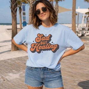 Puede incluir: Camiseta azul claro con el texto "Sad but Healing" en naranja y negro con un estilo retro. Se combina con pantalones cortos de mezclilla. La imagen est&aacute; tomada al aire libre con palmeras y una playa de fondo.