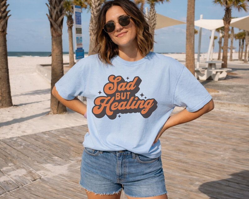 Puede incluir: Camiseta azul claro con el texto "Sad but Healing" en naranja y negro con un estilo retro. Se combina con pantalones cortos de mezclilla. La imagen est&aacute; tomada al aire libre con palmeras y una playa de fondo.
