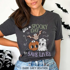 Puede incluir: Camiseta gris oscuro jaspeado con un dise&ntilde;o de Halloween. El dise&ntilde;o presenta un murci&eacute;lago, una calabaza y un fantasma con gorro de enfermera, con el texto "It's a Spooky Day to Save Lives."