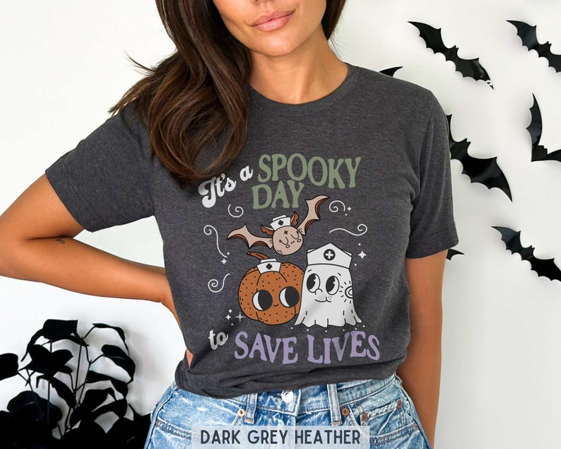 Puede incluir: Camiseta gris oscuro jaspeado con un dise&ntilde;o de Halloween. El dise&ntilde;o presenta un murci&eacute;lago, una calabaza y un fantasma con gorro de enfermera, con el texto "It's a Spooky Day to Save Lives."