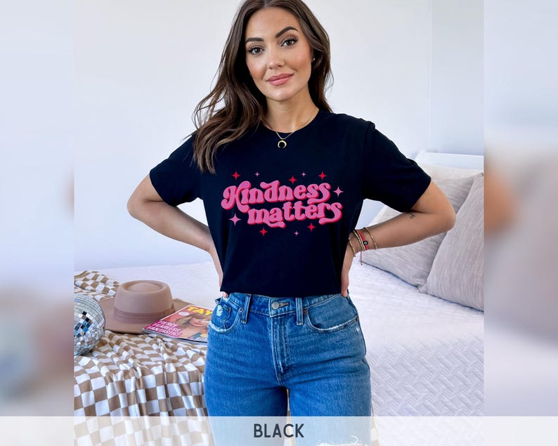 Pode incluir: T-shirt preto com a frase "Kindness matters" em rosa e vermelho. A camisa tem gola redonda e mangas curtas. A modelo est&aacute; usando jeans azuis.