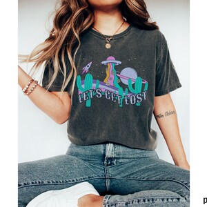 May include: Dark grey t-shirt with a colourful graphic featuring a cactus, a rocket, and a planet, with the text "LET'S GET LOST". The design includes purple, teal, and yellow elements. The shirt has a crew neck.