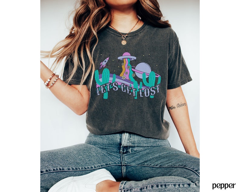 May include: Dark grey t-shirt with a colourful graphic featuring a cactus, a rocket, and a planet, with the text "LET'S GET LOST". The design includes purple, teal, and yellow elements. The shirt has a crew neck.