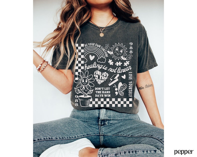 May include: A dark grey t-shirt with a white graphic design featuring a flower, a rainbow, a heart, a lightning bolt, a butterfly, and the text "Be your own sunshine", "healing is not linear", "Find joy in the journey", "Don't let the hard days win", "Love yourself", and "988".
