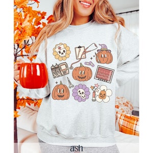 May include: A gray sweatshirt with a Halloween-themed design featuring pumpkins, a skull, a stethoscope, and a heart monitor. The design is in a cartoon style and includes the text "ash".
