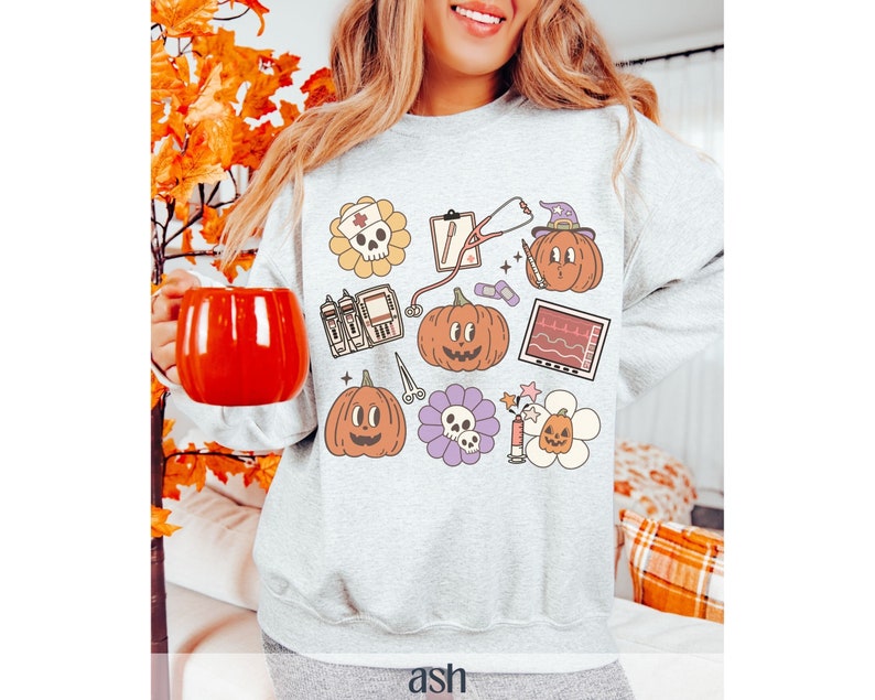 May include: A gray sweatshirt with a Halloween-themed design featuring pumpkins, a skull, a stethoscope, and a heart monitor. The design is in a cartoon style and includes the text "ash".