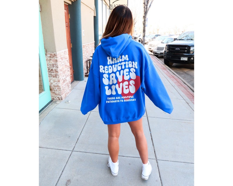 Peut inclure: Un sweat-shirt bleu roi avec le texte blanc "HARM REDUCTION SAVES LIVES" au-dessus d'un c&oelig;ur rouge. En dessous, le texte dit "THERE ARE MULTIPLE PATHWAYS TO RECOVERY". Le sweat-shirt est port&eacute; &agrave; l'ext&eacute;rieur.