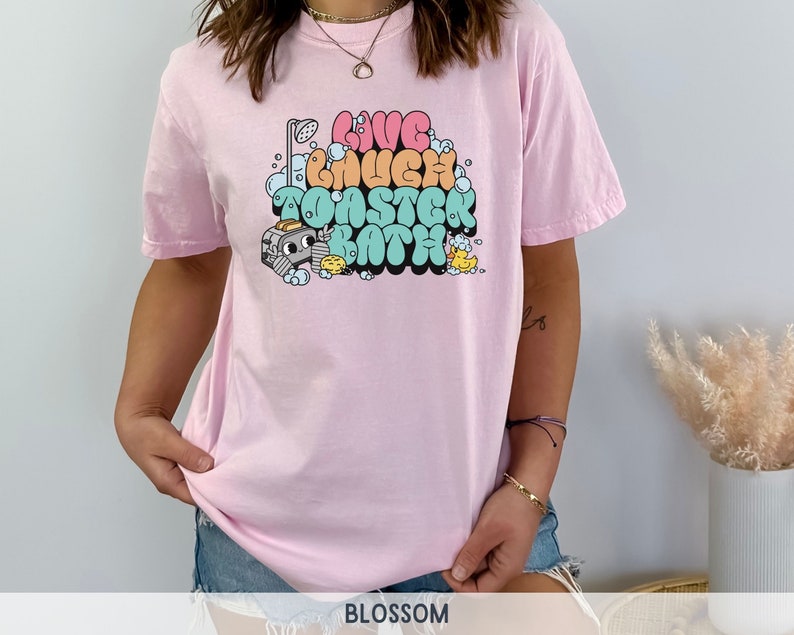 Pu&ograve; includere: Maglietta rosa con una grafica di una doccia di cartoni animati, bolle e il testo "Live Laugh Toaster Bath".