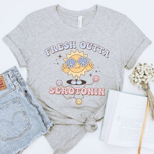 Op de afbeelding: Lichtgrijs t-shirt met de tekst "FRESH OUTTA SEROTONIN" in een retro lettertype. De afbeelding toont een lachende zon met een peace-teken zonnebril. Het shirt wordt gecombineerd met een spijkerbroek en een boek.