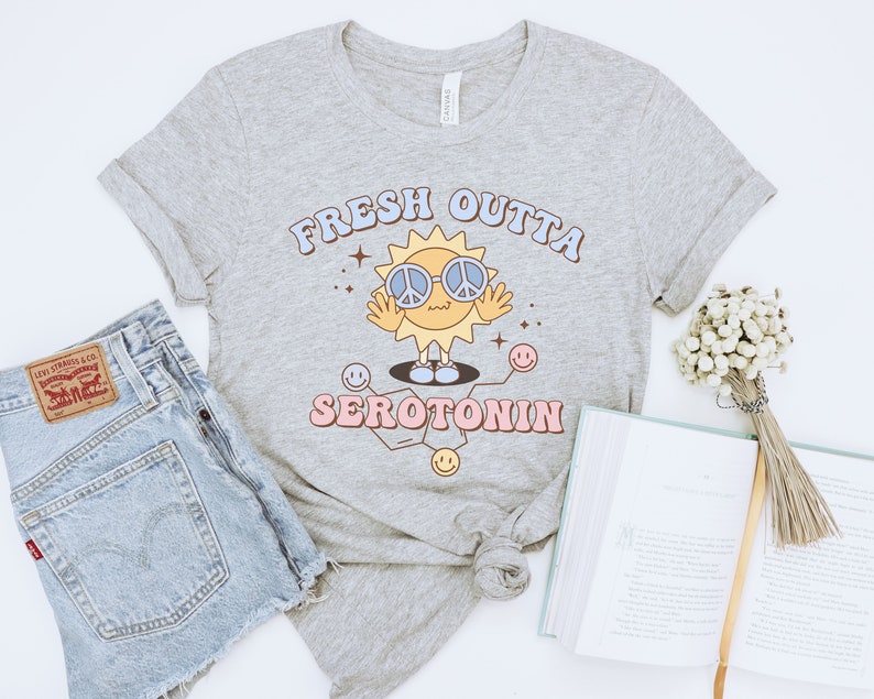 Op de afbeelding: Lichtgrijs t-shirt met de tekst "FRESH OUTTA SEROTONIN" in een retro lettertype. De afbeelding toont een lachende zon met een peace-teken zonnebril. Het shirt wordt gecombineerd met een spijkerbroek en een boek.